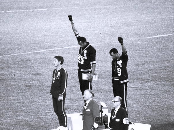 John Carlos et Tommie Smith levant le poing recouvert d'un gant noir sur le podium olympique, accompagnés de Peter Norman